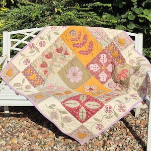 Könnte beinhalten: Ein quadratischer Quilt mit einem Patchwork-Design aus cremefarbenen, senfgelben und dunkelroten Stoffbahnen. Der Quilt zeigt gestickte Blumenmotive, blättrige Ranken und Herzformen, abgerundet durch eine hellviolette Umrandung und feine Nähte.