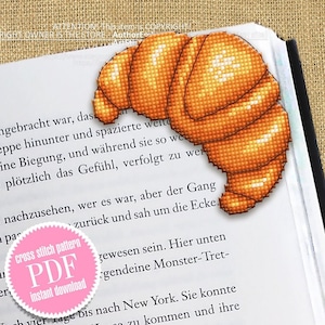 May include: A pink and white digital download button with the text "cross stitch pattern PDF Instant download" on a book page with a crocheted image of a croissant.
