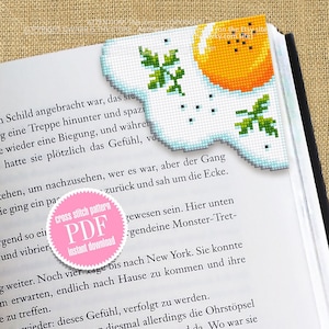May include: A pink circle with the text "cross stitch pattern PDF instant download" in white. The circle is on a book page with text about a story. The book page is folded over and a cross stitch bookmark of a sunny-side-up egg with parsley is visible.