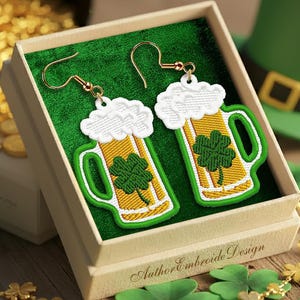 May include: Embroidered St. Patrick's Day earrings in a gift box. The earrings are shaped like beer mugs with green shamrocks and white foam. A green leprechaun hat and gold coins are in the background. The text "Author Embroide Design" is on the box.
