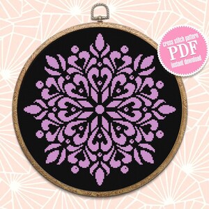May include: A cross-stitch pattern in a wooden hoop. The design is a symmetrical floral pattern in light purple on a black background. A pink sticker with the text "cross stitch pattern PDF instant download" is on the right.