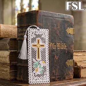 May include: A white rectangular lace bookmark featuring an embroidered gold cross and white lily flowers with green leaves. The bookmark has a scalloped edge and a white tassel attached to the top. The text FSL is visible in the upper right corner.