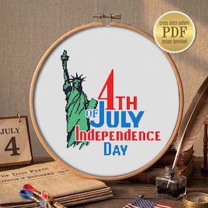 Peut inclure: Un motif de point de croix dans un cerceau en bois représente la Statue de la Liberté et le texte "4th of July Independence Day". Un petit drapeau américain, un calendrier indiquant le 4 juillet et d'autres articles patriotiques sont disposés sur une surface en bois.