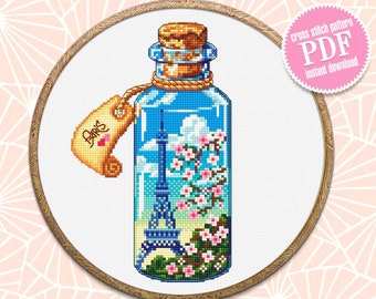 May include: A cross stitch pattern of a glass bottle with a cork stopper. The bottle contains a miniature Eiffel Tower and pink cherry blossoms. A tag attached to the bottle says "Paris" with a red heart.