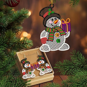 Puede incluir: Adorno navideño y juego de pendientes de muñeco de nieve bordado. El muñeco de nieve tiene un sombrero de copa negro, una bufanda roja y verde y sostiene un regalo morado. Los pendientes se muestran en una caja pequeña.