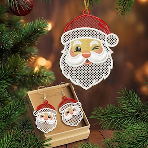Puede incluir: Un conjunto de adorno navideño y pendientes a juego con un diseño de Papá Noel guiñando un ojo. El adorno cuelga de un cordón dorado, mientras que los pendientes se presentan en una pequeña caja. El diseño de Papá Noel tiene un gorro rojo, barba blanca y mejillas rosadas. El texto "2 sizes" es visible.