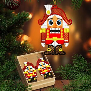 Puede incluir: Imagen navideña con un adorno de cascanueces rojo, blanco y dorado colgado de un árbol de Navidad. Un par de pendientes de cascanueces a juego se muestran en una pequeña caja. El texto "2 tamaños" es visible.