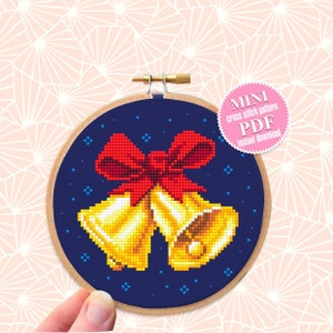 May include: A cross stitch pattern featuring two gold bells with a red bow on a dark blue background with white stars. The pattern is in a round wooden embroidery hoop. The text "MINI cross stitch pattern PDF Instant download" is on a pink circle.