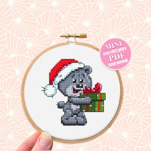 Könnte beinhalten: Ein grauer Teddybär mit einem roten Weihnachtsmann-Hut hält ein grün-rot verpacktes Geschenk. Der Bär befindet sich in einem hölzernen Stickrahmen. Der Text "MINI cross stitch pattern PDF Instant download" befindet sich auf einem rosa Kreis.