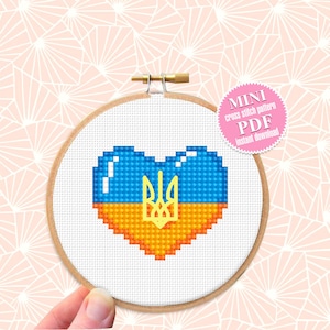 May include: A cross-stitch embroidery hoop featuring a heart design in the colors of the Ukrainian flag: blue and yellow. The Ukrainian coat of arms is in the center. The text "MINI cross stitch pattern PDF instant download" is on a pink sticker.