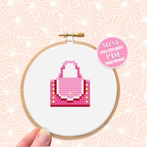May include: A pink cross stitch pattern of a small handbag with a yellow handle and accents. The pattern is displayed in a wooden embroidery hoop. The text "MINI cross stitch pattern PDF Instant download" is on a white circle with a pink border.