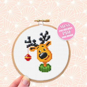 May include: A cross stitch pattern of a cartoon reindeer with a red ornament hanging from its antlers. The pattern is in a round embroidery hoop and is labeled "MINI cross stitch pattern PDF Instant Download".