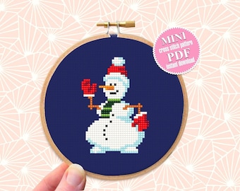 Può includere: Un modello di punto croce di un pupazzo di neve con un cappello rosso, una sciarpa verde e guanti rossi. Il modello è su uno sfondo blu ed è incorniciato in un cerchio di legno. Il testo "MINI cross stitch pattern PDF Instant Download" è su un cerchio rosa.