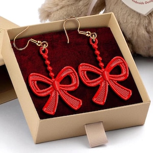 Puede incluir: Pendientes rojos con forma de lazo y ganchos dorados, presentados en una caja beige con interior de terciopelo rojo. Un oso de peluche con una cinta y una etiqueta está en el fondo. La etiqueta dice "AUTHOR Embroidery Design".