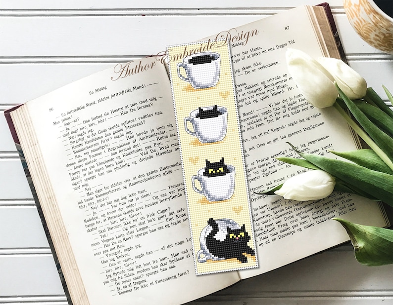Puede incluir: Un marcap&aacute;ginas con fondo amarillo y un gato negro asom&aacute;ndose a una taza de t&eacute;. El marcap&aacute;ginas tiene cuatro im&aacute;genes de gatos en tazas de t&eacute;, cada uno con una postura diferente. El texto "Author Embroide Design" est&aacute; impreso en el marcap&aacute;ginas.