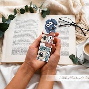 May include: A handmade bookmark with three cartoon dog faces in blue, brown, and grey. The bookmark is held over an open book, with a cup of coffee, glasses, and a candle in the background. The text "Author Embroide Design" is visible.