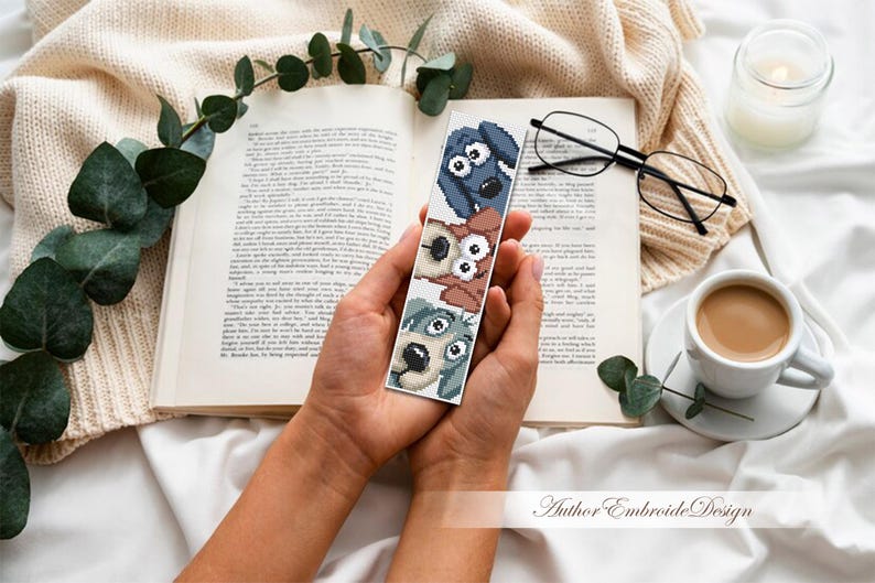 May include: A handmade bookmark with three cartoon dog faces in blue, brown, and grey. The bookmark is held over an open book, with a cup of coffee, glasses, and a candle in the background. The text "Author Embroide Design" is visible.