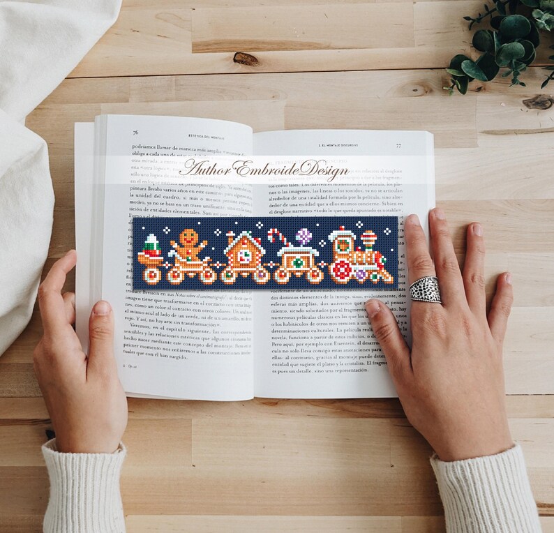 Puede incluir: Un marcap&aacute;ginas bordado a punto de cruz con un dise&ntilde;o de tren navide&ntilde;o, que incluye un hombre de jengibre y una casa. El marcap&aacute;ginas, con el texto "Author Embroide Design", est&aacute; colocado en un libro abierto. El dise&ntilde;o presenta acentos coloridos.