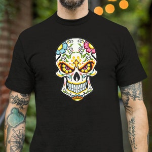 Puede incluir: Una camiseta negra con un dise&ntilde;o de calavera de az&uacute;car colorida. La calavera tiene una cara blanca con ojos negros, una nariz roja y una boca amarilla. La calavera est&aacute; decorada con flores y enredaderas en tonos de rosa, azul, amarillo y verde.