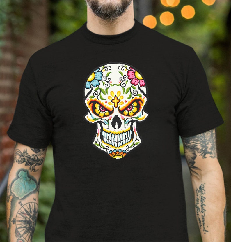 Puede incluir: Una camiseta negra con un dise&ntilde;o de calavera de az&uacute;car colorida. La calavera tiene una cara blanca con ojos negros, una nariz roja y una boca amarilla. La calavera est&aacute; decorada con flores y enredaderas en tonos de rosa, azul, amarillo y verde.