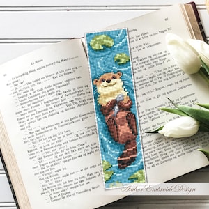 May include: A handmade bookmark featuring a pixelated otter holding a fish, set against a blue water background with lily pads. The bookmark is placed on an open book with white tulips and text in the background. The text "Author Embroide Design" is visible.