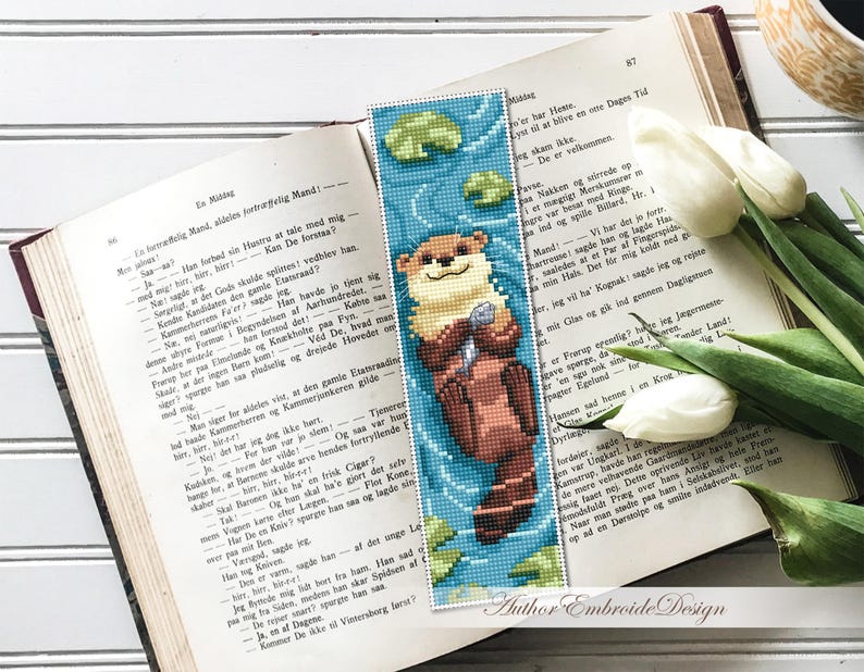 May include: A handmade bookmark featuring a pixelated otter holding a fish, set against a blue water background with lily pads. The bookmark is placed on an open book with white tulips and text in the background. The text "Author Embroide Design" is visible.