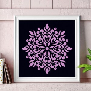 May include: A framed cross-stitch artwork featuring a symmetrical, floral-like design in light purple against a black background. The artwork is displayed in a white frame. The image is of a decorative item.