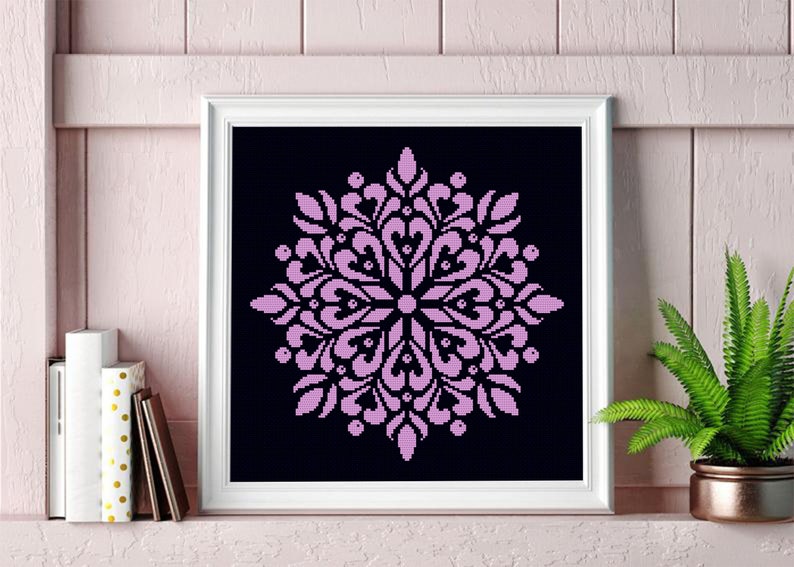 May include: A framed cross-stitch artwork featuring a symmetrical, floral-like design in light purple against a black background. The artwork is displayed in a white frame. The image is of a decorative item.