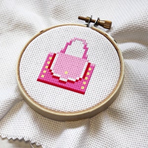 May include: A pink and yellow pixelated handbag cross-stitch pattern on white Aida cloth in a wooden embroidery hoop.