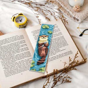 May include: A beaded bookmark featuring an otter holding a shell, set against a blue background with lily pads. The bookmark is placed in an open book, next to a yellow alarm clock, glasses, and a cup of tea. The scene is on a white surface.