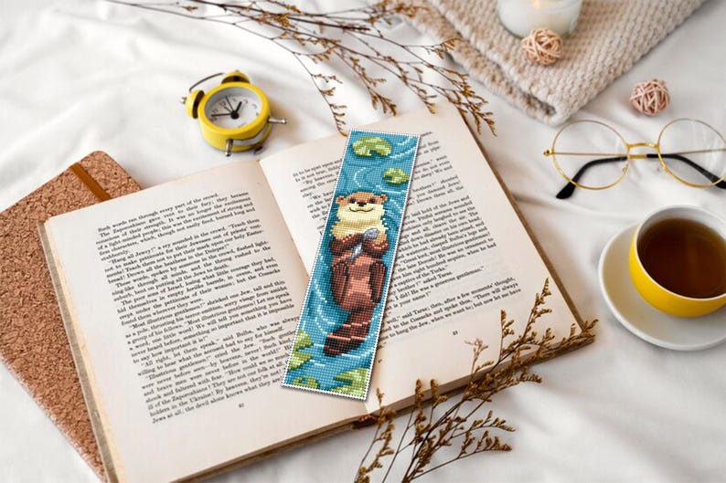 May include: A beaded bookmark featuring an otter holding a shell, set against a blue background with lily pads. The bookmark is placed in an open book, next to a yellow alarm clock, glasses, and a cup of tea. The scene is on a white surface.