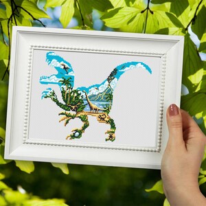 May include: A framed cross-stitch design featuring a blue dinosaur silhouette with a tropical island scene inside. The dinosaur is outlined in black and the island scene includes palm trees, a beach, and a blue sky.