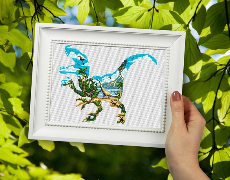 May include: A framed cross-stitch design featuring a blue dinosaur silhouette with a tropical island scene inside. The dinosaur is outlined in black and the island scene includes palm trees, a beach, and a blue sky.