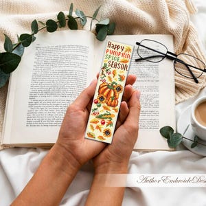 May include: A decorative bookmark with the text "Happy Pumpkin Spice Season" and autumn-themed illustrations of pumpkins, sunflowers, and leaves. The bookmark is held over an open book, with a cup of coffee and glasses nearby.
