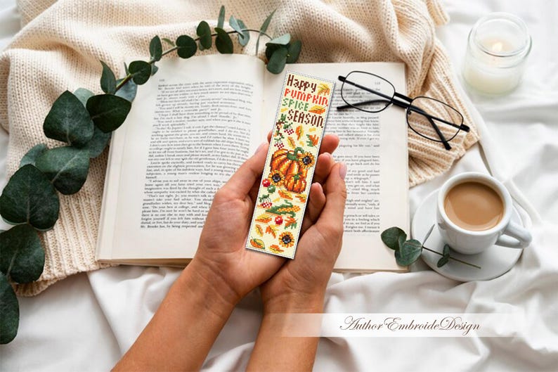 May include: A decorative bookmark with the text "Happy Pumpkin Spice Season" and autumn-themed illustrations of pumpkins, sunflowers, and leaves. The bookmark is held over an open book, with a cup of coffee and glasses nearby.