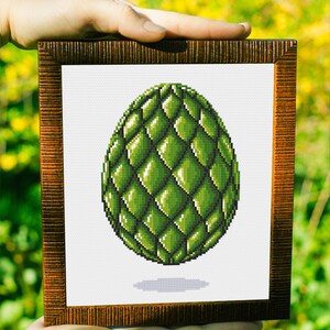 May include: A framed cross-stitch art piece featuring a green, scaled egg design. The egg is shaded with varying tones of green, creating a three-dimensional effect. The frame is a dark brown color with a textured surface. The background is blurred.