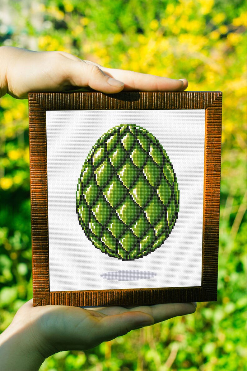 May include: A framed cross-stitch art piece featuring a green, scaled egg design. The egg is shaded with varying tones of green, creating a three-dimensional effect. The frame is a dark brown color with a textured surface. The background is blurred.