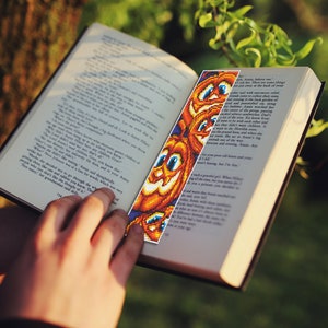 May include: A close-up of a book with a colorful beaded bookmark featuring a pattern of orange pumpkins with smiling faces. The bookmark is made of small beads and is a fun and festive way to mark your place in a book.