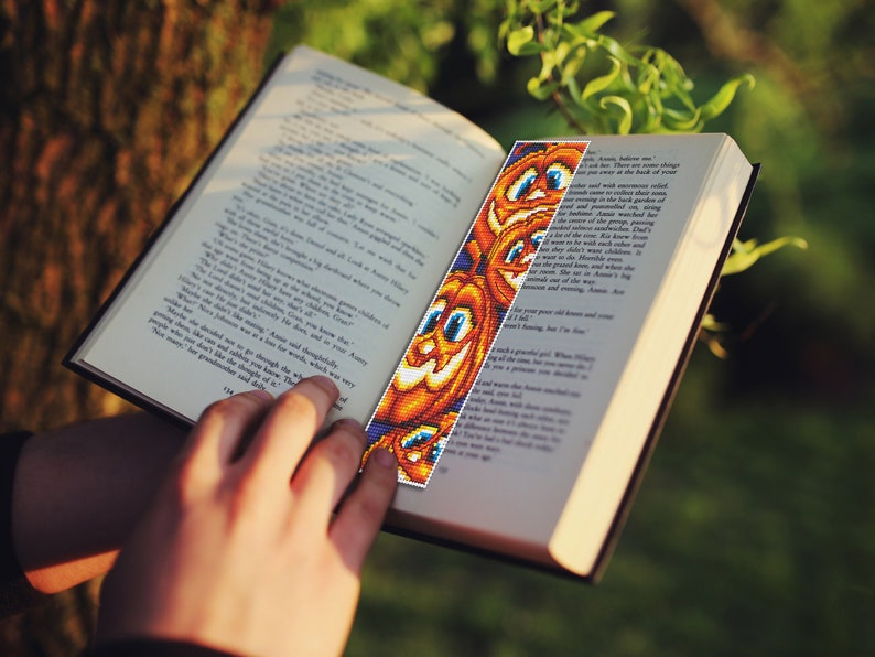 May include: A close-up of a book with a colorful beaded bookmark featuring a pattern of orange pumpkins with smiling faces. The bookmark is made of small beads and is a fun and festive way to mark your place in a book.
