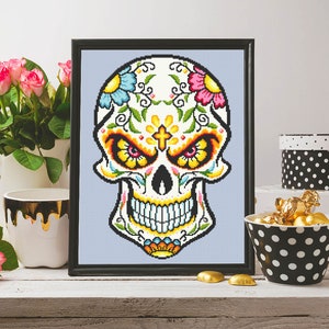 Puede incluir: Un dise&ntilde;o de punto de cruz enmarcado de una calavera de az&uacute;car blanca con flores coloridas y una cruz en la frente. La calavera sonr&iacute;e con ojos amarillos y un dise&ntilde;o detallado.