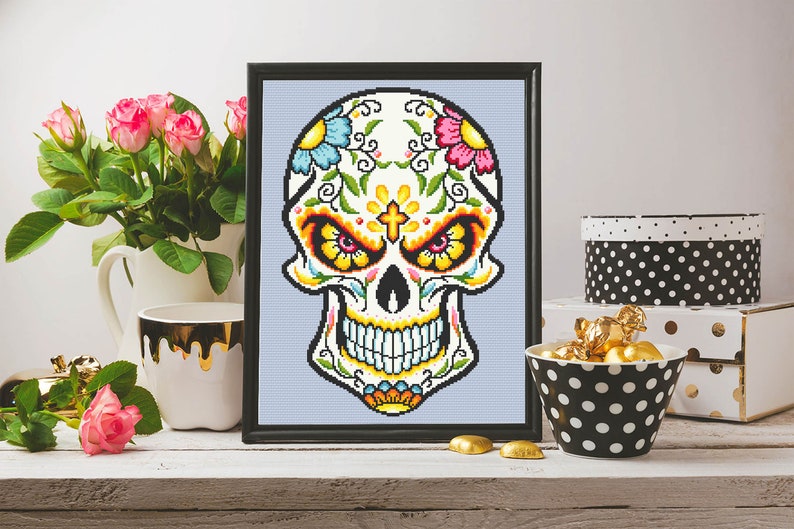 Puede incluir: Un dise&ntilde;o de punto de cruz enmarcado de una calavera de az&uacute;car blanca con flores coloridas y una cruz en la frente. La calavera sonr&iacute;e con ojos amarillos y un dise&ntilde;o detallado.