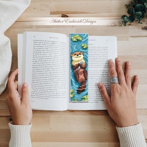 May include: A handmade bookmark featuring a pixelated otter design. The otter is brown and beige, floating in blue water with lily pads. The bookmark is placed in an open book. The text "Author Embroide Design" is at the top.