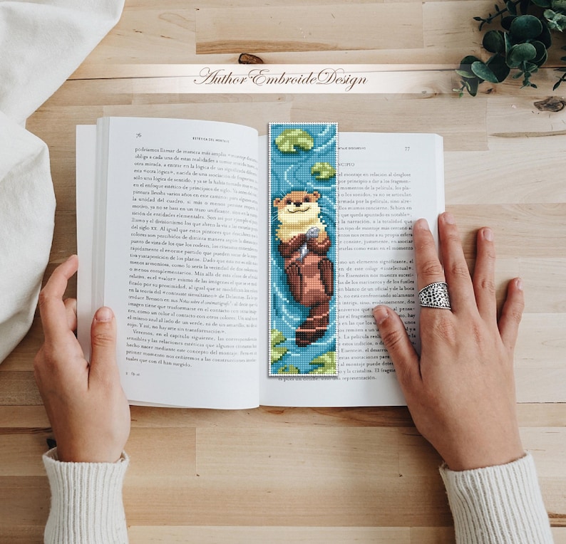 May include: A handmade bookmark featuring a pixelated otter design. The otter is brown and beige, floating in blue water with lily pads. The bookmark is placed in an open book. The text "Author Embroide Design" is at the top.