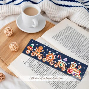 Puede incluir: Un marcap&aacute;ginas azul marino con un dise&ntilde;o pixelado de tren navide&ntilde;o con un hombre de jengibre, una casa y un bast&oacute;n de caramelo. El marcap&aacute;ginas est&aacute; sobre un libro abierto con una taza de caf&eacute; cerca.