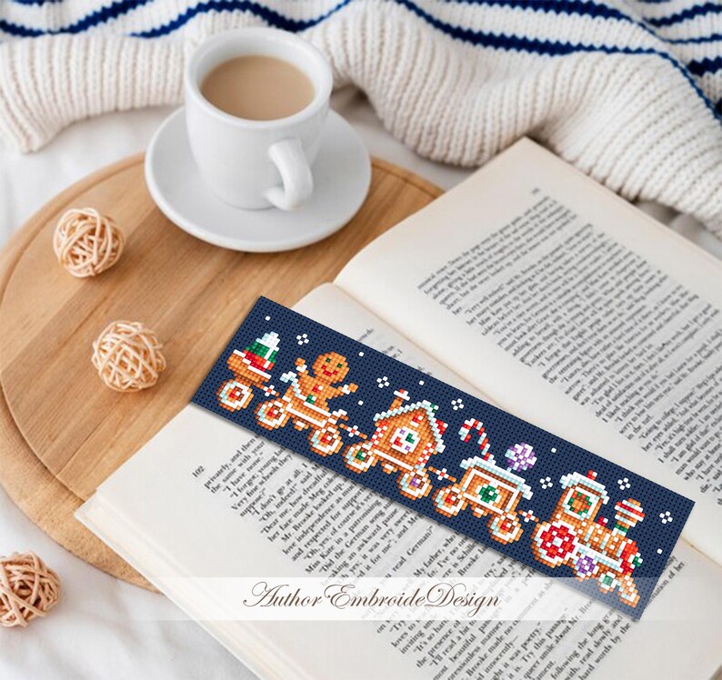 Puede incluir: Un marcap&aacute;ginas azul marino con un dise&ntilde;o pixelado de tren navide&ntilde;o con un hombre de jengibre, una casa y un bast&oacute;n de caramelo. El marcap&aacute;ginas est&aacute; sobre un libro abierto con una taza de caf&eacute; cerca.