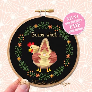 May include: A black and white cross stitch pattern of a chicken with the text "Guess what..." in a wreath of green leaves and orange flowers. The pattern is in a wooden embroidery hoop.
