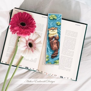 May include: A handmade bookmark featuring a pixelated otter holding a shell, set against a blue water background with lily pads. The bookmark is placed in an open book next to pink and white gerbera daisies.