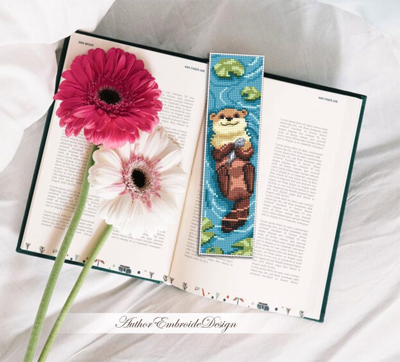 May include: A handmade bookmark featuring a pixelated otter holding a shell, set against a blue water background with lily pads. The bookmark is placed in an open book next to pink and white gerbera daisies.