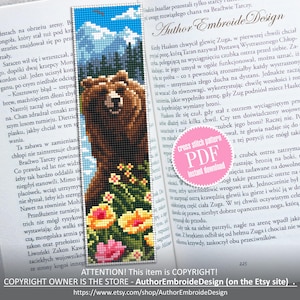 May include: A handmade bookmark featuring a pixelated image of a brown bear in a mountain landscape with colorful flowers. The bookmark is made with cross-stitch technique and is placed in an open book.