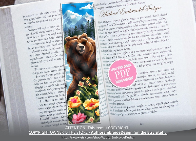 May include: A handmade bookmark featuring a pixelated image of a brown bear in a mountain landscape with colorful flowers. The bookmark is made with cross-stitch technique and is placed in an open book.